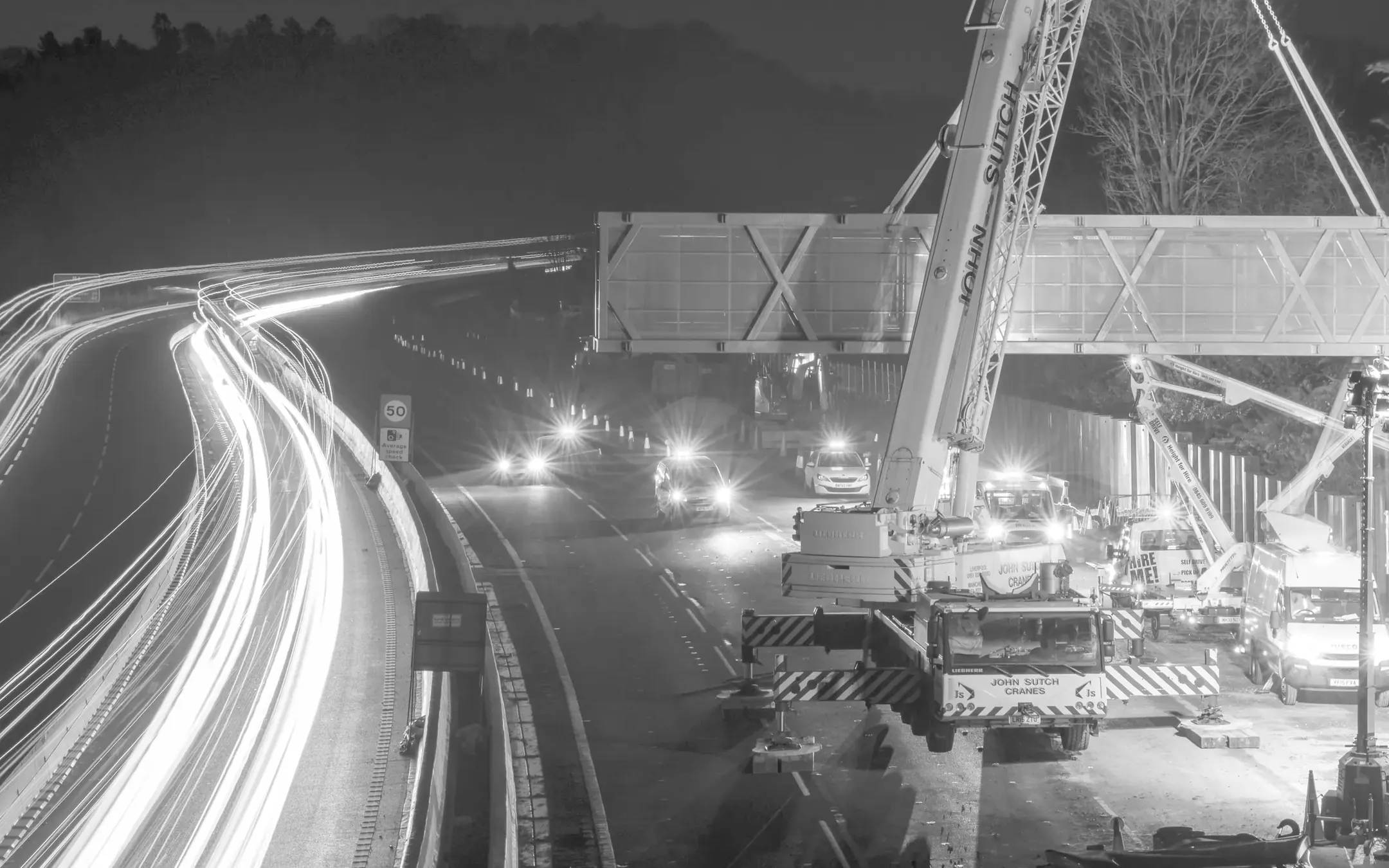 Traffic light trails past a gantry being lifted over a motorway at night